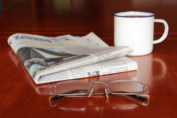 Zeitung, Lesebrille und Tasse Kaffee auf Holztisch
