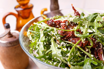 Green healthy salad with arugula and sun-dried tomatoes and parmesan cheese on a wooden kitchen table. Pepper, peppermoll, utensils for olive oil, vinegar and seasonings. Italian cuisine