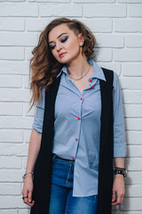 Beautiful and sassy woman in blue shirt, casual long vest and blue jeans posing against a white brick wall. Eyes closed.