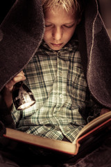 Child reading book under covers with flashlight