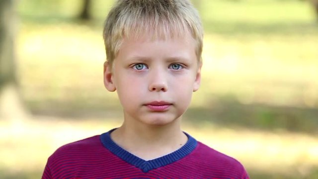 Close up portrait of angry 8 years old boy over green trees background. Angry funny face of little kid and his fist up. Child lokking at camera isolated at nature background. Real time video footage