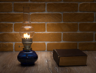 The book by the light of an old lamp. Glass oil lamp and book on the wooden table against the background of a brick wall.