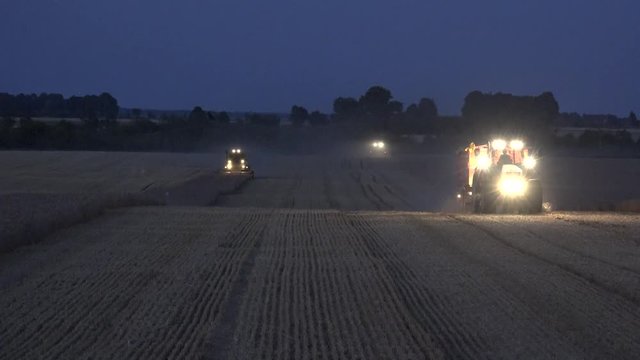 Tractor with trailer and combines with strong light cultivating barley rye wheat field at night. Static shot. 4K 
