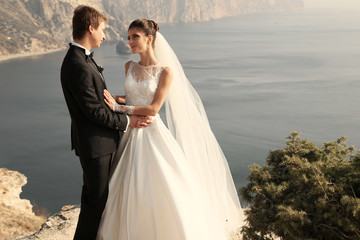 fashion outdoor photo of beautiful couple. gorgeous bride in wedding dress posing with elegant groom on sea cost  