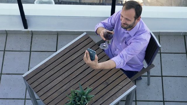 Young man with glass of wine taking selfie photo with cellphone on terrace, top view
