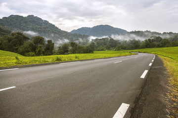 The asphalt way which has been accompanied by mountains and trees. The cloudy sky makes the sky dark.
