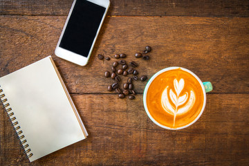 Top view of latte hot coffee, white smart phone and blank note book in coffee shop. Wooden table background.