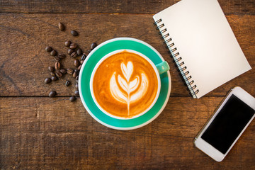 Top view of latte hot coffee, white smart phone and blank note book in coffee shop. Wooden table background.