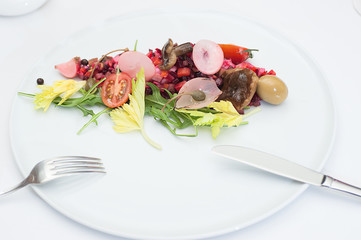 Vinaigrette Russian Salad made of chopped beets, carrots, onion, beans, peas, seasoned with sunflower seed oil, with slices of tomatoes, herbs and sausage, served on a white table with fork and knife