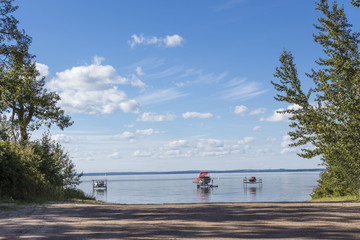 horizontal image of two boats and a sea doo  docked  close to the shore line framed by two tall green trees under a beautiful blue sky in the summer time.