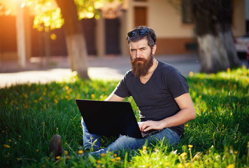 brutal bearded man with a mustache T-shirt, torn jeans and shoes, sitting with a laptop in the hands in the spring park on sunset