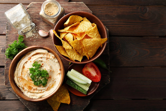 Wooden bowl of tasty hummus with chips, parsley on table