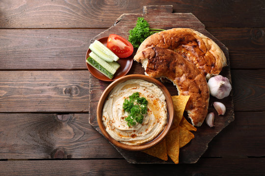 Wooden bowl of tasty hummus with chips, flat bread and parsley on table