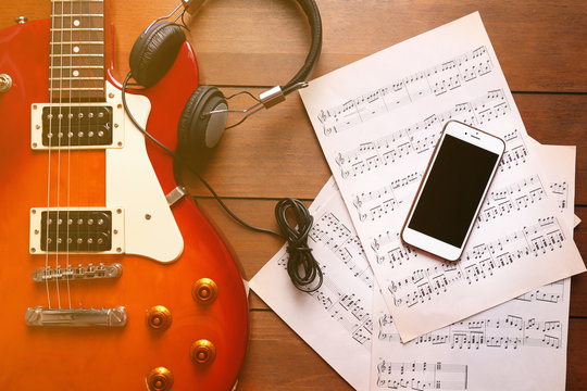 Electric guitar with headphones, mobile phone and notes on wooden background