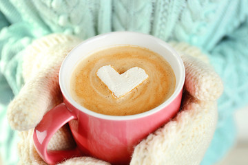 Female hands in warm mittens holding cup of hot cappuccino with heart marshmallow, close up