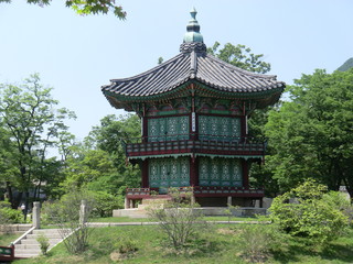 A two storey wood built Pagoda style building in Seoul, South Korea.  Grass and bushes in the foreground, trees in the background with a clear blue sky.