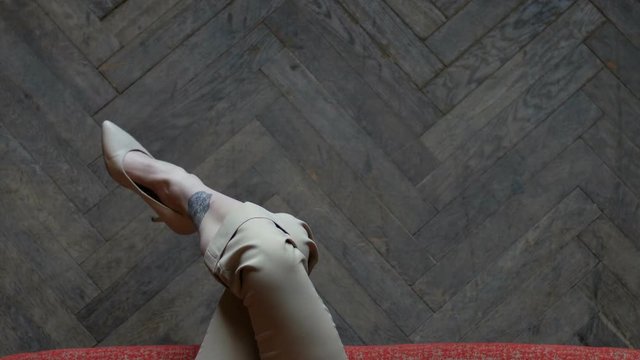Long slim tattoed legs crossed in beige stylish heels. Female legs on a wooden parquet background.