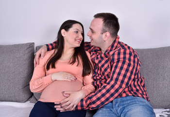 Young smiling couple in love expecting a baby. They are sitting on the sofa and enjoying.