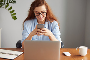 Portrait of beautiful female secretary wearing blue shirt and glasses, smiling while texting on the mobile phone. Young businesswoman typing on the cell phone while sitting in front of the computer