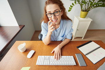Obraz premium Top shot view of young attractive woman smiling and touching her lips while sitting at the wooden table in front of the computer screen during working day. Human face expressions and emotions