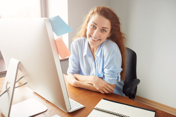 Fototapeta na wymiar Confident attractive female entrepreneur looking and smiling at the camera. Portrait of happy successful businesswoman with red hair and in blue shirt sitting crossed arms in front of the computer