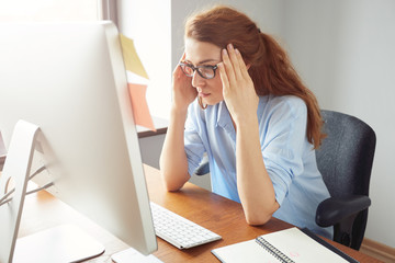 Confident woman freelancer sitting in front of the computer with serious and thoughtful expression. Young female entrepreneur in blue shirt and glasses using PC for work sitting in the office