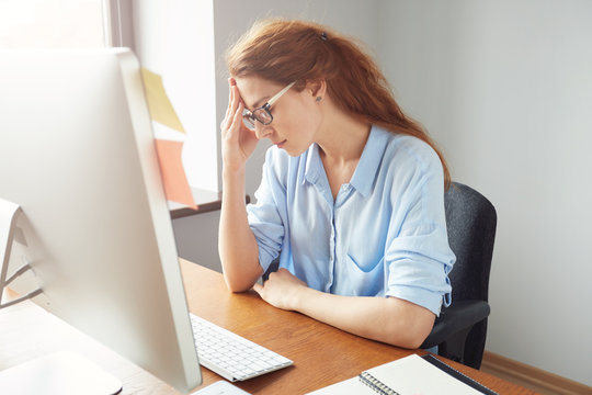 Concerned Female Entrepreneur Looking At The Computer Screen While Working In The Office. Stressed Young Woman Sitting At The Desk And Leaning On Her Elbow While Thinking On Something Important