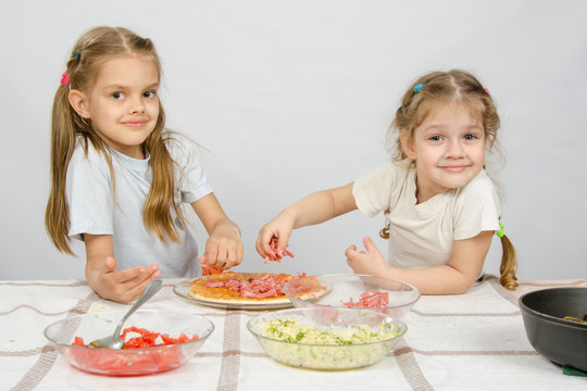 Two little happy girl at the table spread on the pizza ingredients