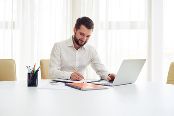 Smiling man working on his laptop and writing some data in a not