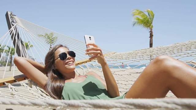 Happy beach woman reading sms texting on smartphone lying on hammock relaxing on tropical vacation. Young casual cute girl lying down in outdoor swing bed enjoying sun sunbathing using mobile phone.