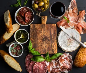 Fototapeten Wine snack set with empty wooden board in center. Glass of red, meat selection, mediterranean olives, sun-dried tomatoes, baguette slices, camembert cheese and spices on black background, © sonyakamoz