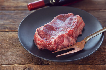 Raw pork steak with meat fork and bottle of red wine on black plate