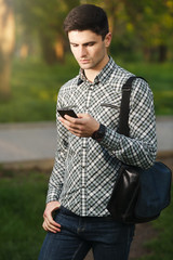 Handsome young man looking at the smart phone, standing in the park area.