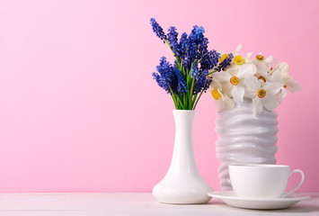 a bouquet of white narcissi in a white vase near the beautiful blue flowers in a vase on a wooden table beside a Cup of coffee on a pink background