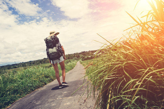 Traveling Woman With Backpack And Hat Walking Along The Road At Sunny Day (intentional Sun Glare)