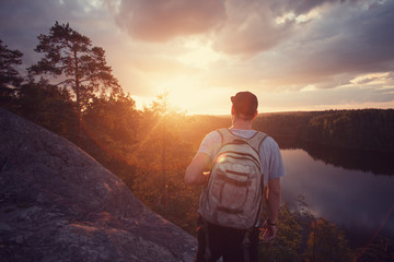 Young casual man on the cliff above river and looking far away at sunset (intentional sun glare)