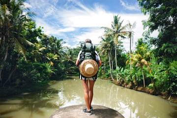Woman with backpack standing on the edge near big tropical river and looking far away