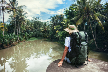 Woman traveler with backpack sitting on the edge near tropical river 
