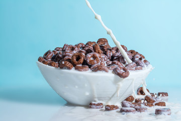 Milk stream jet pouring into a bowl with chocolate flakes in the form of rings, splashes of milk on a light blue celestial background
