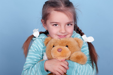 Smiling kid girl 3-4 year old playing with teddy bear over blue. Wearing blue striped hoodie. 