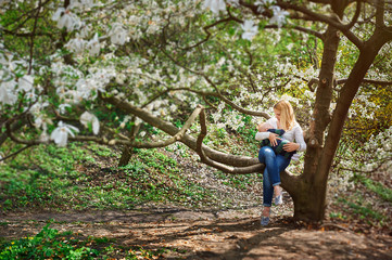 Naklejka na ściany i meble Mom with baby boy sitting on tree branch in the flowered spring garden