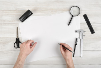 Closeup hands of man holding pencil and drawing on white paper in top view. Draftsman workplace equipped with ruler, pen, stapler, scissors, magnifying glass.