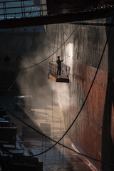 Shipyard worker power washing a ship on dry dock.