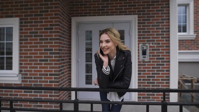 Beautiful girl in leather jacket near its entrance. She is talking to someone on the phone and smiling. 