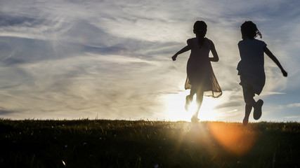 Silhouettes of two young children running towards the sunset - obrazy, fototapety, plakaty