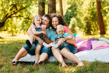 Fototapeta premium Young happy family with kids having picnic with colored pillows outdoors. Parents with two children relax in a sunny summer garden. Mother, father, little girl and baby boy playing in park.