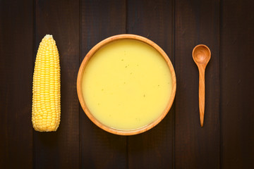 Cream of corn soup in wooden bowl with corn cob and wooden spoon on the side, photographed on dark...