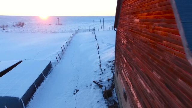 Slow aerial flyby of a red barn shining brilliantly at sunset.  The area around is covered with fresh snow.  Filmed on a family farm in Kansas, USA.