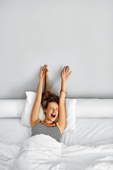 Morning Wake Up. Smiling Young Woman Waking Up Fully Rested On White Bedding. Model Stretching In Bed. Girl Lying, Relaxing In Bedroom. Healthy Sleep, Lifestyle. Wellness, Health, Beauty Concept