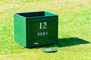 Circular marker lying on tee on a golf course. Nicely cut grass surround it. Peg box for broken pegs on hole 12, a par 4 hole.